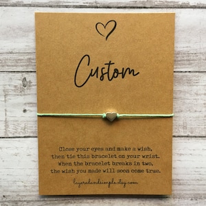 Puede incluir: Una tarjeta marrón con la palabra "Custom" escrita en cursiva negra. Una pulsera de cuerda verde claro con un dije de corazón plateado está atada alrededor de la tarjeta. La tarjeta tiene el texto "Cierra los ojos y pide un deseo, luego ata esta pulsera en tu muñeca. Cuando la pulsera se rompa en dos, el deseo que hiciste pronto se hará realidad. layeredandsimple.etsy.com"