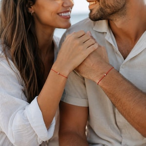 May include: Two matching minimalist bracelets, each consisting of a thin red cord featuring a single small, round, metallic silver bead centered on the wrist. The bracelets are worn on the wrists of two individuals dressed in light-colored clothing.