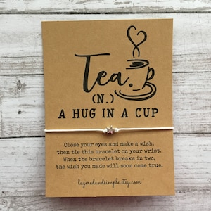 May include: A brown card with the text "Tea (N.) A hug in a cup" and a black illustration of a teacup with steam. A white string bracelet with a silver star charm is attached to the card. The text "Close your eyes and make a wish, then tie this bracelet on your wrist. When the bracelet breaks in two, the wish you made will soon come true." is printed on the card. The website layeredandsimple.etsy.com is printed at the bottom of the card.
