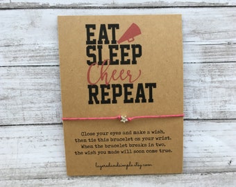 Peut inclure: Une carte marron avec le texte "EAT SLEEP Cheer REPEAT" et un bracelet en cordon rose avec un charme étoile argenté. La carte contient également des instructions pour faire un vœu sur le bracelet.