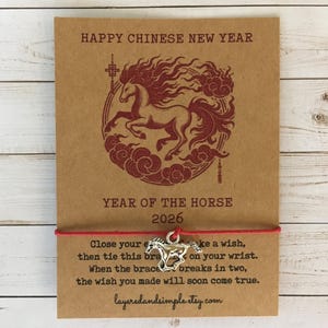 Pode incluir: Um cartão castanho com texto vermelho e uma ilustração de cavalo vermelho, com uma pulseira de corda vermelha e um berloque de cavalo prateado. O texto diz "Happy Chinese New Year" e "Year of the Horse 2026".