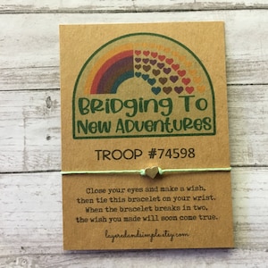 May include: A green and white string bracelet with a gold heart charm. The bracelet is attached to a card with a rainbow and the text "Bridging To New Adventures" and "TROOP #74598". The card also includes instructions for making a wish with the bracelet.