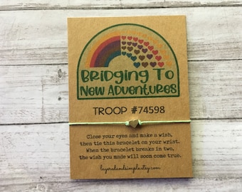 May include: A green and white string bracelet with a gold heart charm. The bracelet is attached to a card with a rainbow and the text "Bridging To New Adventures" and "TROOP #74598". The card also includes instructions for making a wish with the bracelet.