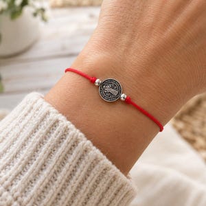 May include: A red string bracelet featuring a circular silver-toned metal charm with an embossed religious figure and Latin text. The charm is flanked by two small silver-toned beads on either side, secured by a thin, vibrant red cord band.