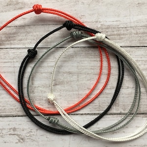 May include: Four bracelets made of thin cord in red, black, gray, and white. The bracelets are tied in a simple knot.