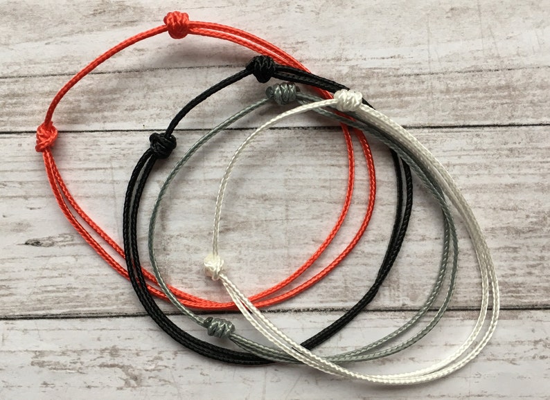 May include: Four bracelets made of thin cord in red, black, gray, and white. The bracelets are tied in a simple knot.