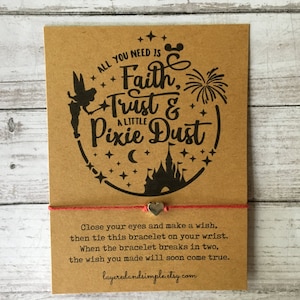 Könnte beinhalten: Eine braune Karte mit einer Schwarzweißillustration einer Fee, Feuerwerk, einem Schloss und Sternen. Der Text lautet "Alles, was du brauchst, ist Glaube, Vertrauen und ein bisschen Feenstaub". Die Karte hat eine rote Schnur, die an einem silbernen Herzanhänger befestigt ist.