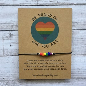 May include: A card with the text "BE PROUD OF WHO YOU ARE" and a rainbow heart design. Below is a black string bracelet with rainbow beads. The card also includes instructions and the Etsy shop URL.