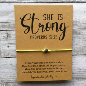 May include: A kraft paper card with the words "SHE IS Strong" and "PROVERBS 31:25" printed in black. A neon green string bracelet with a small silver star charm is attached to the card. The card also includes instructions and a website address.