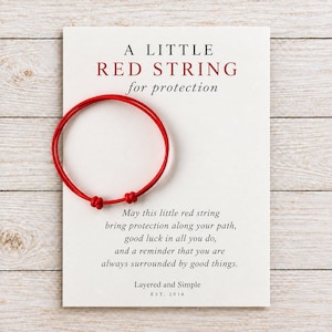 May include: A thin red string bracelet with two adjustable knots, displayed on a white card featuring the text A LITTLE RED STRING for protection, followed by a message about protection and good luck, signed by Layered and Simple EST. 2016.