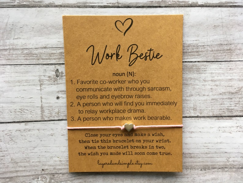 May include: A brown card with the text "Work Bestie" and a definition of the word. The card is tied with a pink string and two gold heart charms.