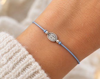 May include: A light blue cord bracelet featuring a small, oval-shaped silver-toned charm depicting a figure in relief. The charm is flanked by two tiny silver-toned beads, one on each side, secured by knots on the thin, smooth cord.