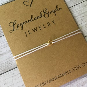 Puede incluir: Una pulsera minimalista con un dije de anillo dorado y cordón blanco. La pulsera se muestra en una tarjeta marrón con el texto "Layered and Simple Jewelry" y la dirección del sitio web. El fondo es de madera clara.