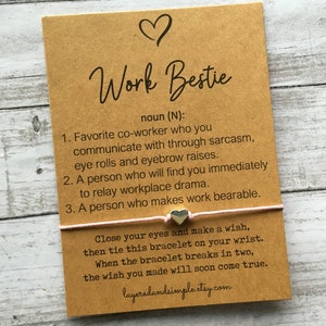 May include: A brown card with the text "Work Bestie" and a definition of the term. The card also includes a pink string bracelet with a silver heart charm and instructions for making a wish.
