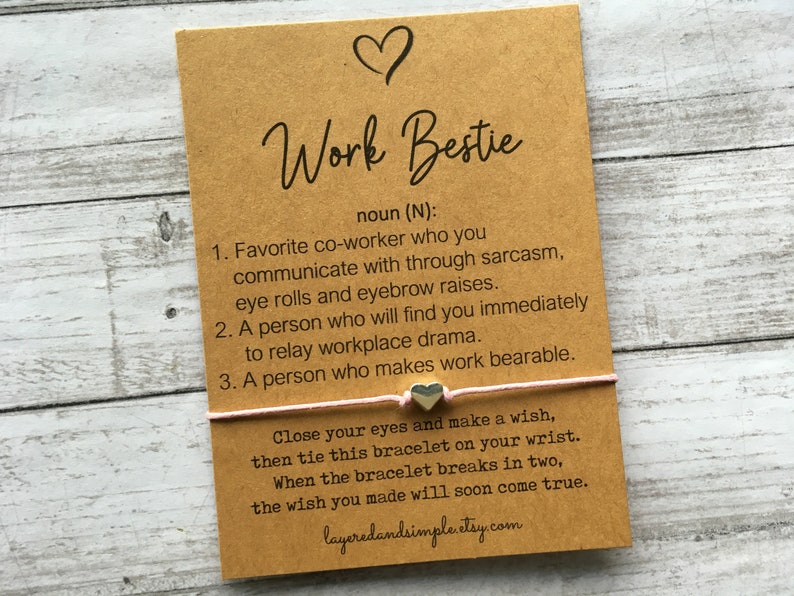 May include: A brown card with the text "Work Bestie" and a definition of the term. The card also includes a pink string bracelet with a silver heart charm and instructions for making a wish.