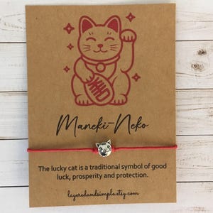 Pode incluir: Pulseira de fio vermelho com um berloque de gato prateado, exibida em um cartão marrom com uma ilustração vermelha de Maneki-Neko. O cartão inclui texto sobre o símbolo do gato da sorte e um endereço web.