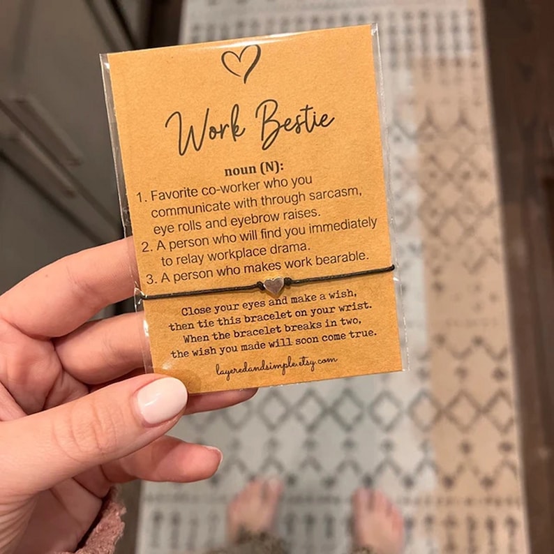 May include: A brown card with the words "Work Bestie" in black lettering. The card defines "Work Bestie" as a co-worker who communicates through sarcasm, eye rolls, and eyebrow raises. The card also includes a brown string bracelet with two silver heart charms.