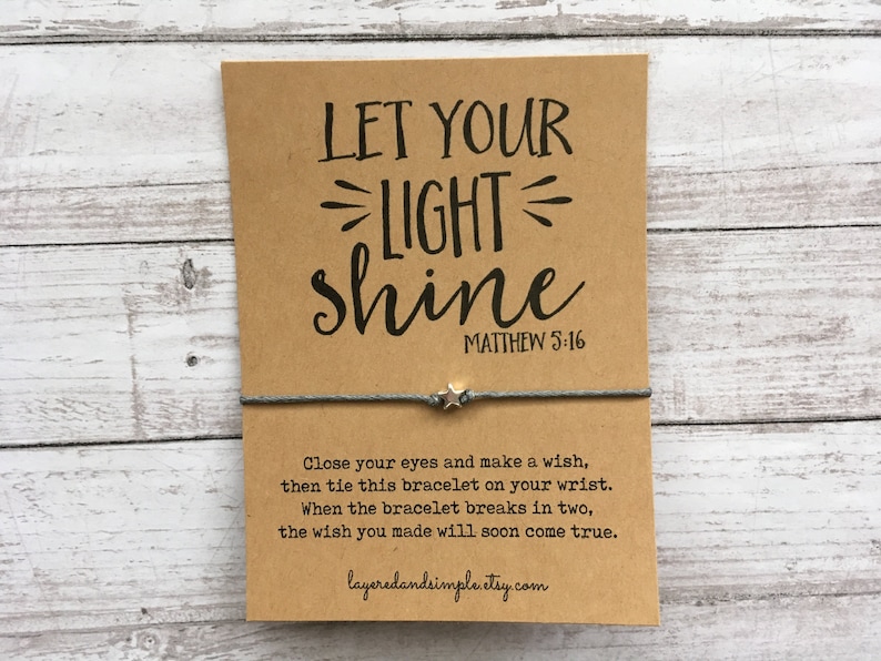 May include: A brown card with the text "LET YOUR LIGHT shine" and "MATTHEW 5:16" printed on it. A gray string bracelet with a silver star charm is tied around the card. The card also includes instructions for making a wish and tying the bracelet.