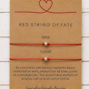 May include: Two red string bracelets with a single silver-toned bead on each, mounted on a rectangular kraft paper card. The card features a red heart illustration, the text RED STRING OF FATE, MINE, YOURS, and a printed quote about destiny.