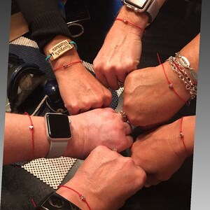 May include: Five people wearing red string bracelets with small silver stars on them. One bracelet has a wooden charm with the text "Seward's  Wisdom".