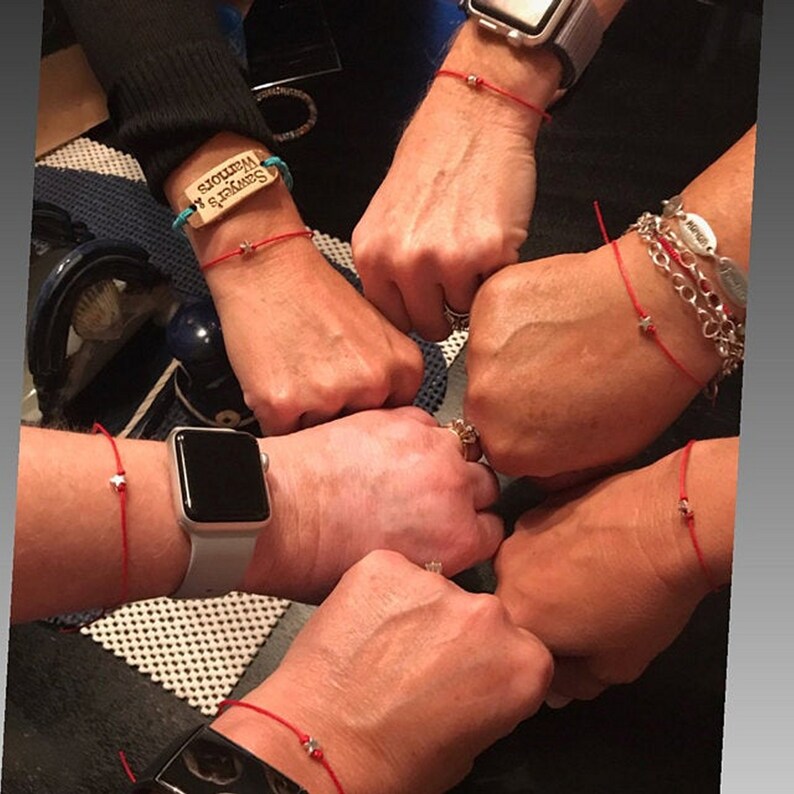 May include: Five people wearing red string bracelets with small silver stars on them. One bracelet has a wooden charm with the text "Seward's  Wisdom".