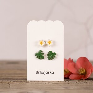 May include: A set of four earrings displayed on a white card with the word "Brlogarka" printed on it. The earrings are made from polymer clay and feature white flowers with yellow centres and green monstera leaves.