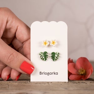 May include: Earrings in the shape of flowers and leaves on a white card. The earrings are white and yellow flowers and green and white monstera leaves. The card has the word "Brlogarka" printed on it.