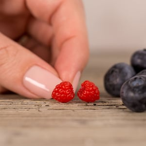 May include: Two red raspberries on a wooden surface, with a hand reaching towards them. There are also several blueberries in the background.