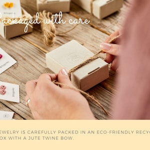 May include: A person carefully ties a jute twine bow around a small, eco-friendly recycled gift box. The box is on a wooden surface, surrounded by other boxes and tags with the text "Briogorko". The words "Packaged with care" are written in cursive.