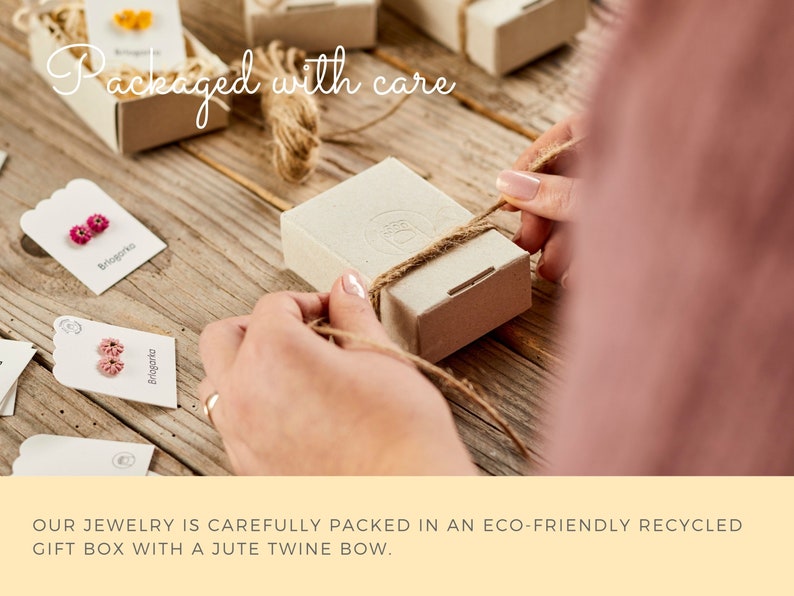 May include: A person carefully ties a jute twine bow around a small, eco-friendly recycled gift box. The box is on a wooden surface, surrounded by other boxes and tags with the text "Briogorko". The words "Packaged with care" are written in cursive.
