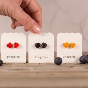 May include: Three pairs of fruit-shaped stud earrings in red, black, and orange, displayed on white cards with the text "Brlogarka". Blueberries are scattered around the earrings on a wooden surface.