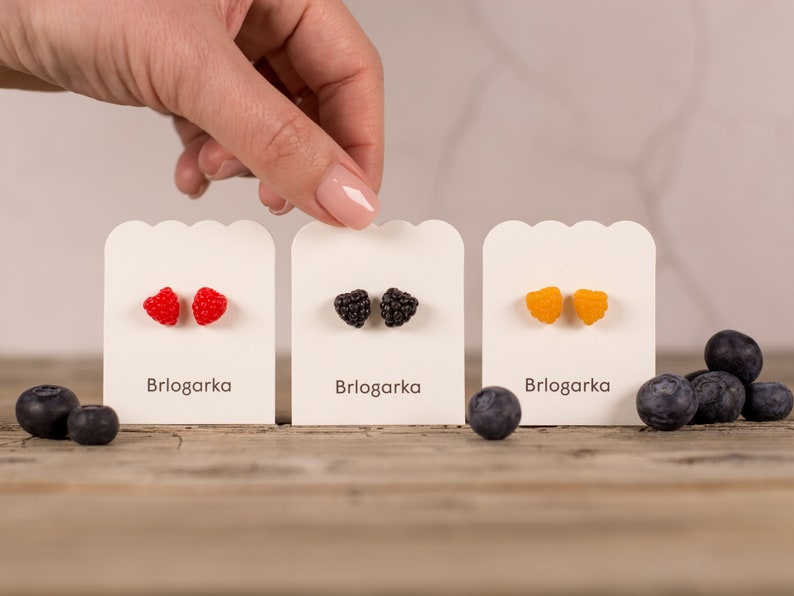 May include: Three pairs of fruit-shaped stud earrings in red, black, and orange, displayed on white cards with the text "Brlogarka". Blueberries are scattered around the earrings on a wooden surface.