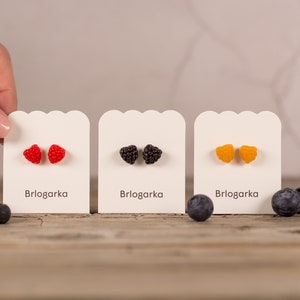May include: Three pairs of fruit-shaped stud earrings on display. The earrings are red raspberries, black blackberries, and yellow raspberries. Each pair is on a white card with the brand name "Brlogarka". Blueberries are scattered around the cards.