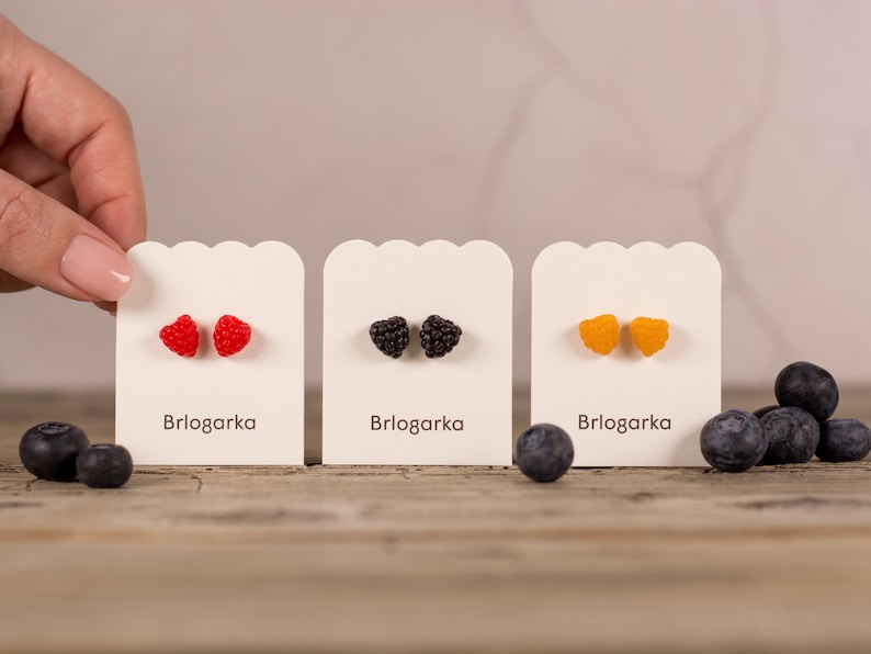 May include: Three pairs of fruit-shaped stud earrings on display. The earrings are red raspberries, black blackberries, and yellow raspberries. Each pair is on a white card with the brand name "Brlogarka". Blueberries are scattered around the cards.
