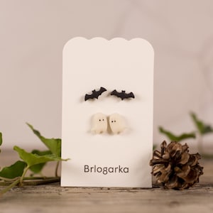 Op de afbeelding: Een witte kaart toont twee paar Halloween-oorbellen: zwarte vleermuizen en witte geesten. Het woord "Brlogarka" staat onderaan gedrukt. Een dennenappel en groen zijn op de voorgrond.