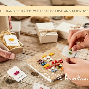 May include: A collection of colorful, handmade earrings displayed on a wooden surface. The image shows various floral and fruit designs, with some earrings packaged in small boxes. Text on the image reads "ALL HAND SCULPTED. WITH LOTS OF LOVE AND ATTENTION." and "100% handmade."