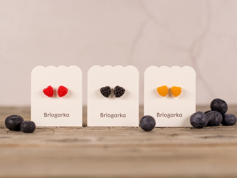 May include: Three pairs of berry-shaped stud earrings on display cards. The earrings are red raspberries, black blackberries, and yellow raspberries. The brand name "Brlogarka" is printed on each card. Blueberries are scattered around the cards.