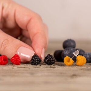May include: A collection of berry-themed earrings and blueberries on a wooden surface. The earrings are designed to resemble raspberries, blackberries, and yellow raspberries. The image is well-lit, showcasing the vibrant colors of the berries.