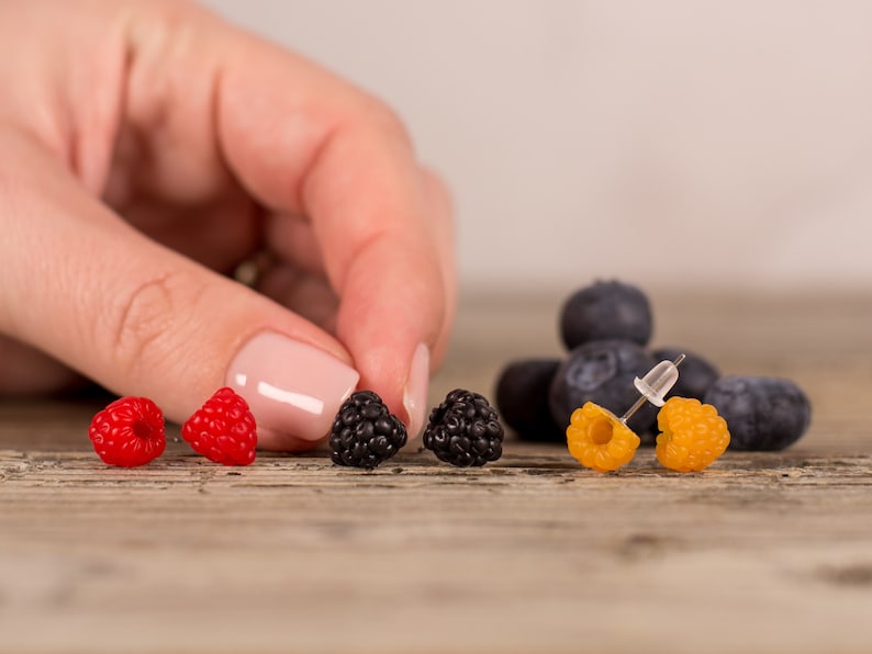 May include: A collection of berry-themed earrings and blueberries on a wooden surface. The earrings are designed to resemble raspberries, blackberries, and yellow raspberries. The image is well-lit, showcasing the vibrant colors of the berries.