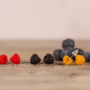 May include: A collection of berry-themed earrings. The earrings are designed to resemble raspberries, blackberries, and golden raspberries. Blueberries are in the background. The earrings are on a wooden surface.