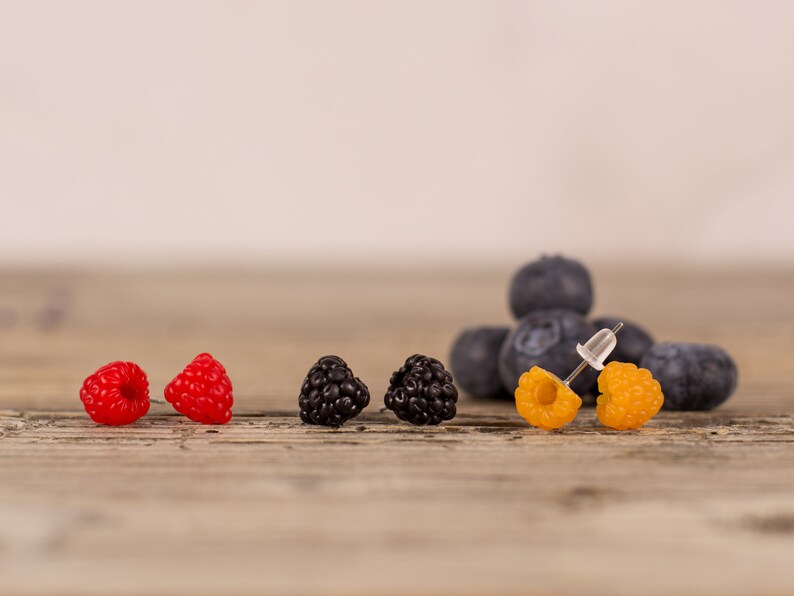 May include: A collection of berry-themed earrings. The earrings are designed to resemble raspberries, blackberries, and golden raspberries. Blueberries are in the background. The earrings are on a wooden surface.