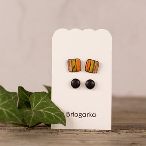 May include: A pair of colorful square earrings with a heart design and black stud earrings on a white card with the text "Brlogarka".