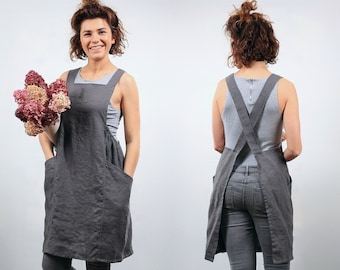 May include: A gray linen apron with two large pockets and a criss-cross back strap. The apron is worn by a person holding a bouquet of pink flowers.