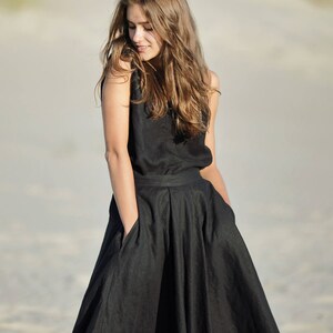 May include: A sleeveless black dress with a full skirt and pockets. The dress is made of a dark fabric and is worn outdoors. The model has long, light brown hair and is looking down.