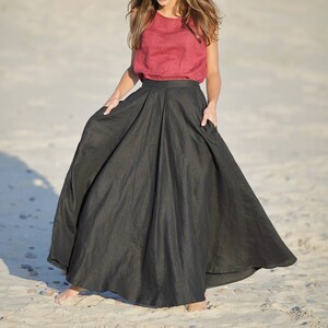 May include: A woman wearing a sleeveless, dark red top and a long, flowing black skirt. The skirt has a high waist and pockets. The outfit is made of a lightweight fabric, suitable for warm weather. The model is standing on a sandy beach.