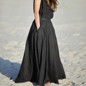 May include: A sleeveless black maxi dress with a wide skirt and pockets. The dress is made of a flowing fabric and is worn outdoors on a sandy beach. The model is wearing the dress.