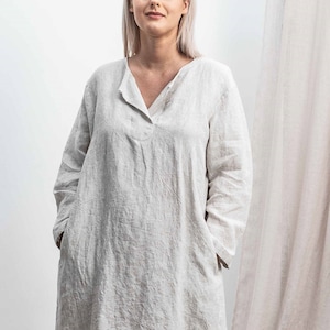 May include: A woman wearing a long, loose-fitting linen dress with a V-neck and long sleeves. The dress is a light beige color and has a textured, linen look. The woman is standing in front of a white background.