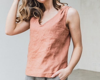 May include: A sleeveless, coral-colored linen top with a V-neck. The top is paired with gray and white striped pants. The model is standing in front of a white wall.