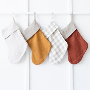 Op de afbeelding: Vier kerstsokken van linnen hangen aan een witte stang. De sokken zijn in verschillende kleuren, waaronder beige, mosterdgeel, beige ruit en roest.