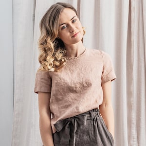May include: A woman wearing a light brown linen top with a brown linen skirt and a brown belt. She is standing in front of a white curtain.